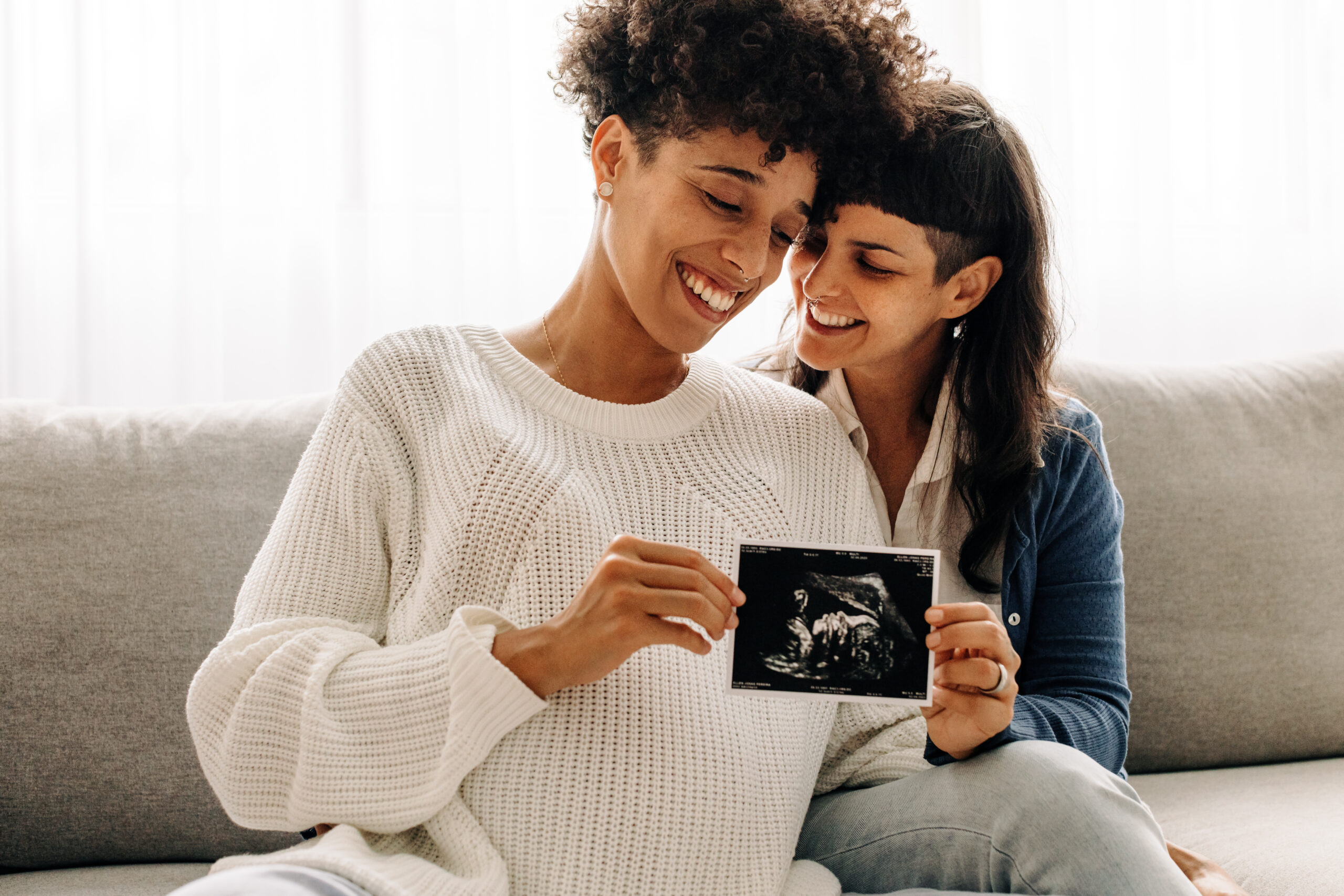 Same-sex pregnant couple holding up their ultrasound scan. Expectant lesbian couple smiling cheerfully while holding an ultrasound picture of their unborn baby. Young queer couple expecting a baby.