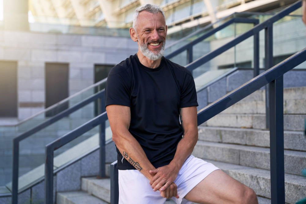 Man wearing shorts and a tshirt stands on outdoor steps, leaning against a barrier and facing the camera.