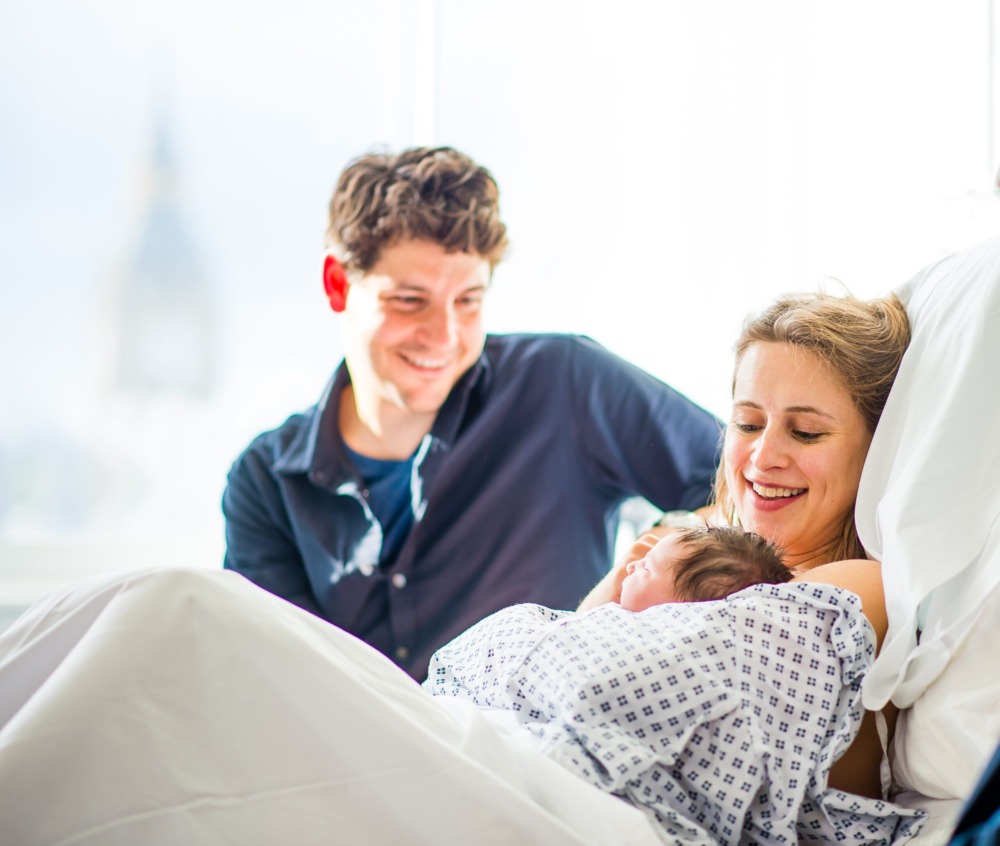 Patient resting in a hospital bed in the Westminster Maternity Suite with a newborn baby and a support person seated nearby in a bright clinical room