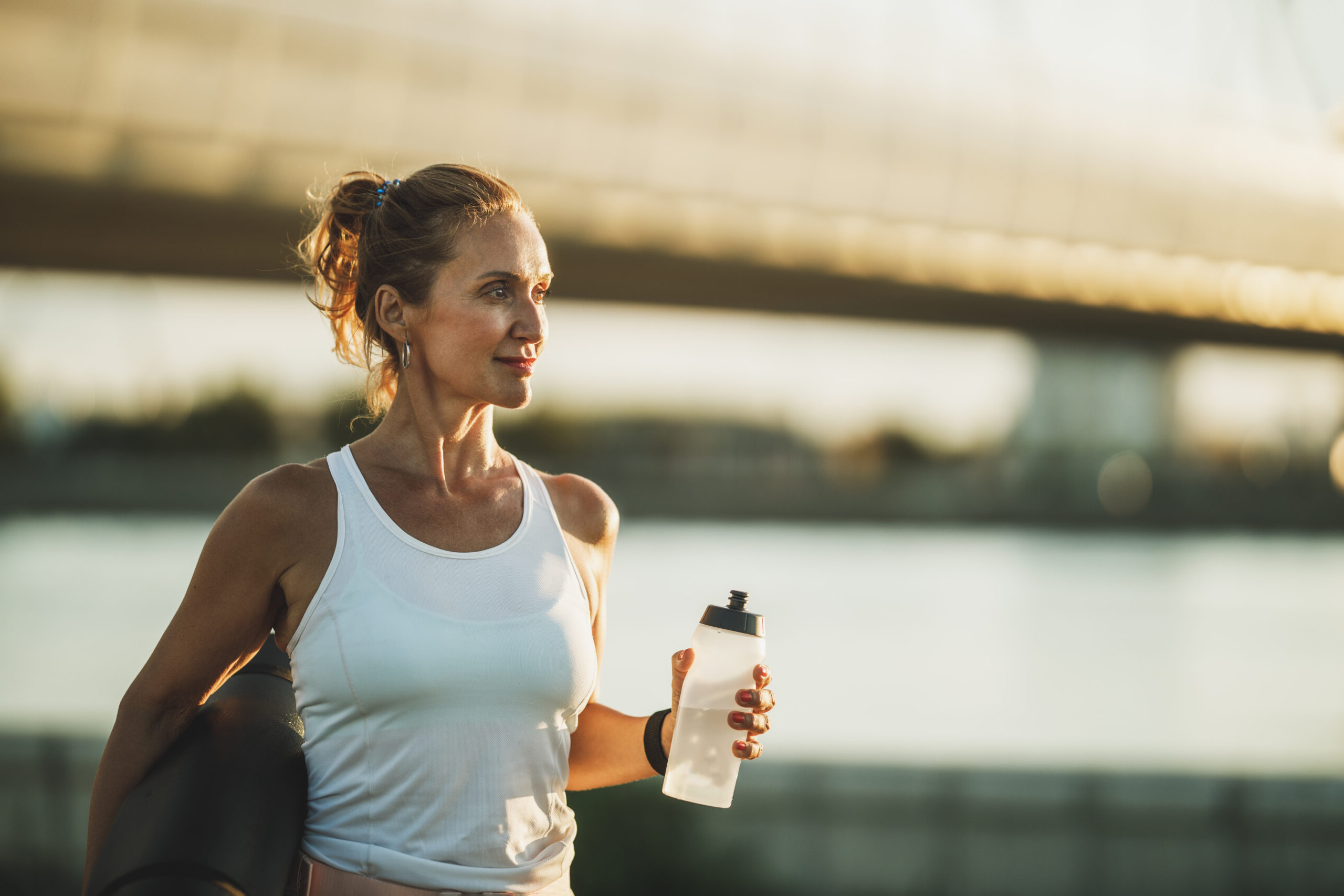 Middle age sportswoman holding water bottle and resting after hard training near to river, in the city.