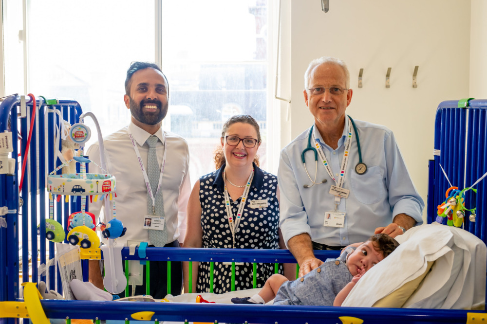 Hospital cot in a clinical ward setting with an infant resting inside, surrounded by medical staff wearing identification lanyards and clinical equipment positioned nearby.