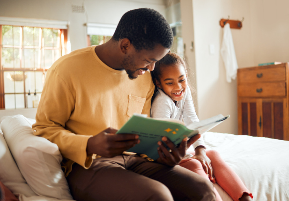 Father sat on the side of a bed with his daughter, reading a storybook to her.