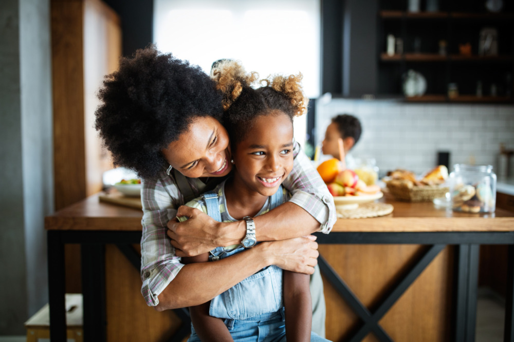 Happy mother hugging her daughter from behind in their kitchen at home.
