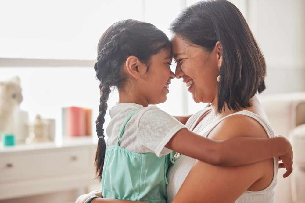 A mother with her daughter sat on her lap. They face each other, smiling and holding each other, with their foreheads touching.