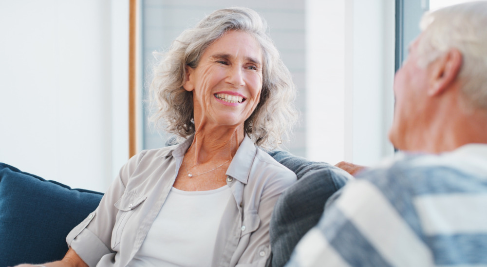 A mature woman on her sofa, smiling widely and in conversation with her husband, who has his back to the camera.