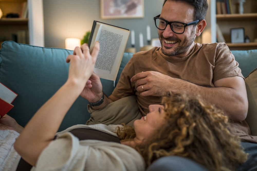 An adult couple laugh together on a sofa. The woman has her head on her partner's lap as she reads a book. He looks at the pages, too.