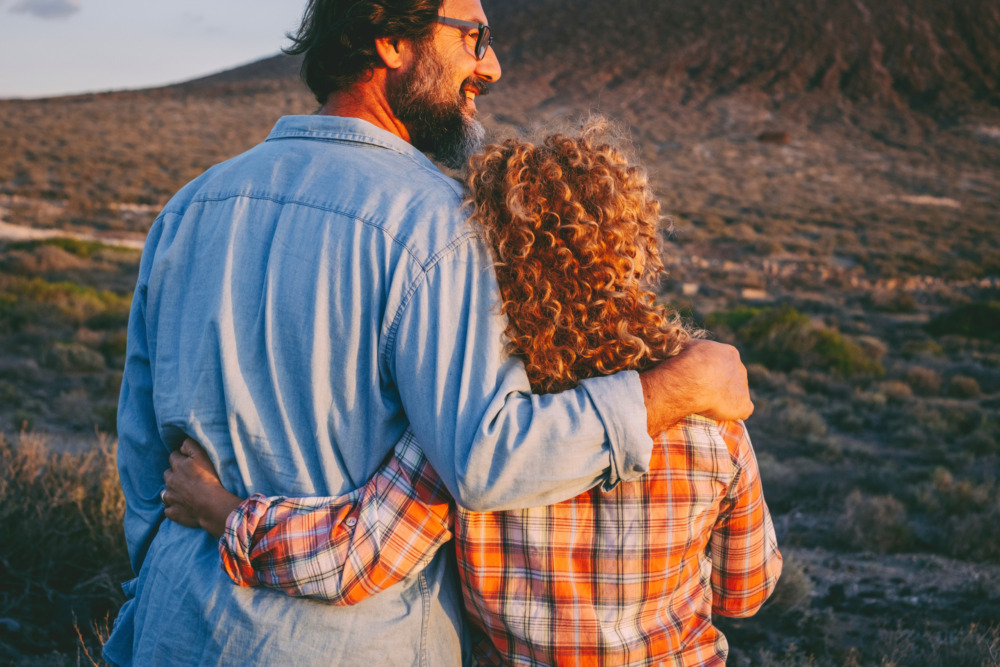 View from behind of a happy couple embracing each other at sunset, admiring scenic country side.