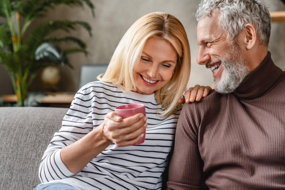 Woman drinking a coffee, leaning on her husband. They are sat on the sofa together.