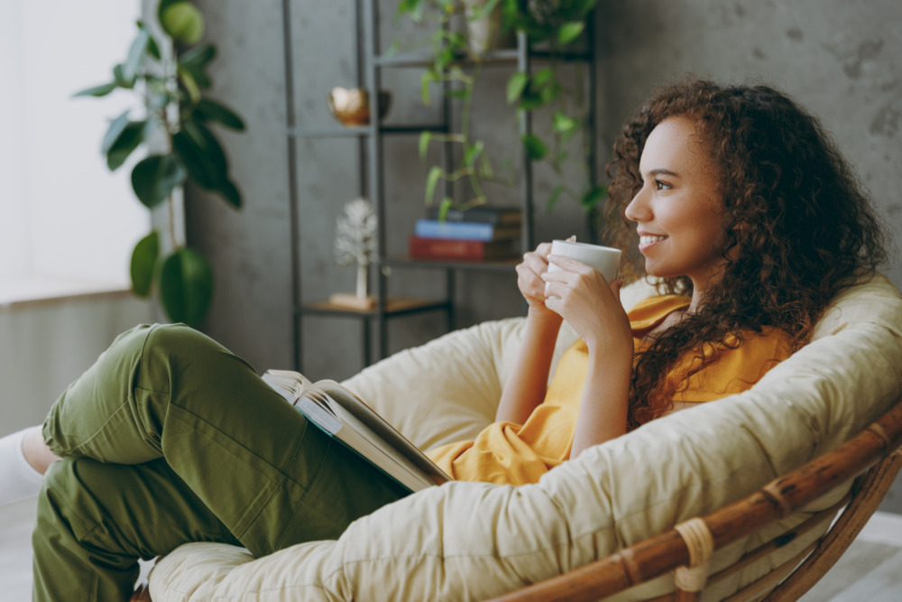 Young woman reading a book and drinking coffee in a comfortable chair, smiling.