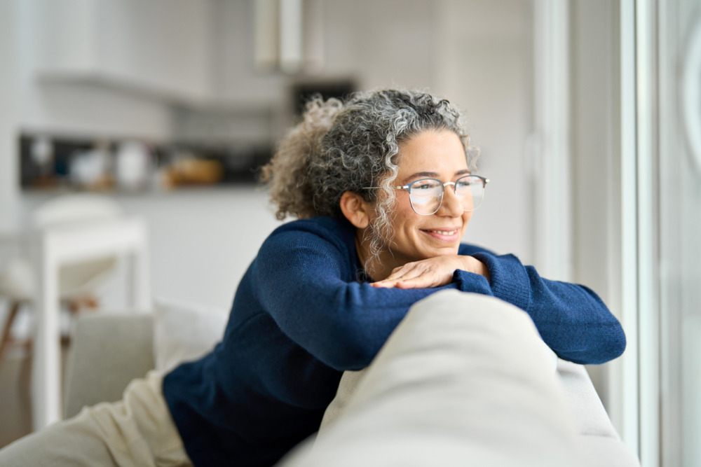 Happy middle aged woman sitting on comfy sofa in living room at home looking through window.