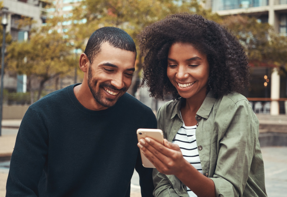 Close-up of a smiling young man looking at mobile phone held by a woman next to him