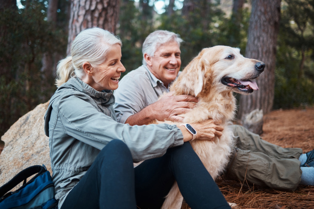 An older couple, hiking, sit either side of their golden retriever, who they both pet as they smile.