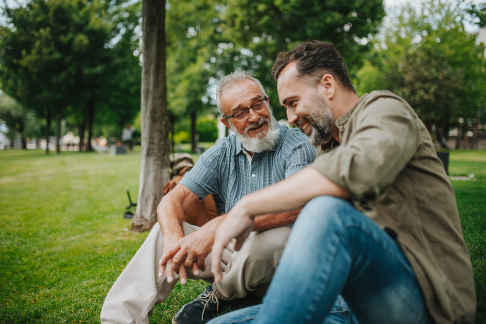 Father and adult son sitting on grass in park, talking and laughing.