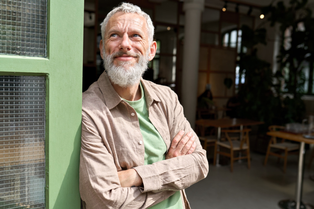 Smiling middle aged man confidently leaning against a wall in the sunshine with his arms folded.