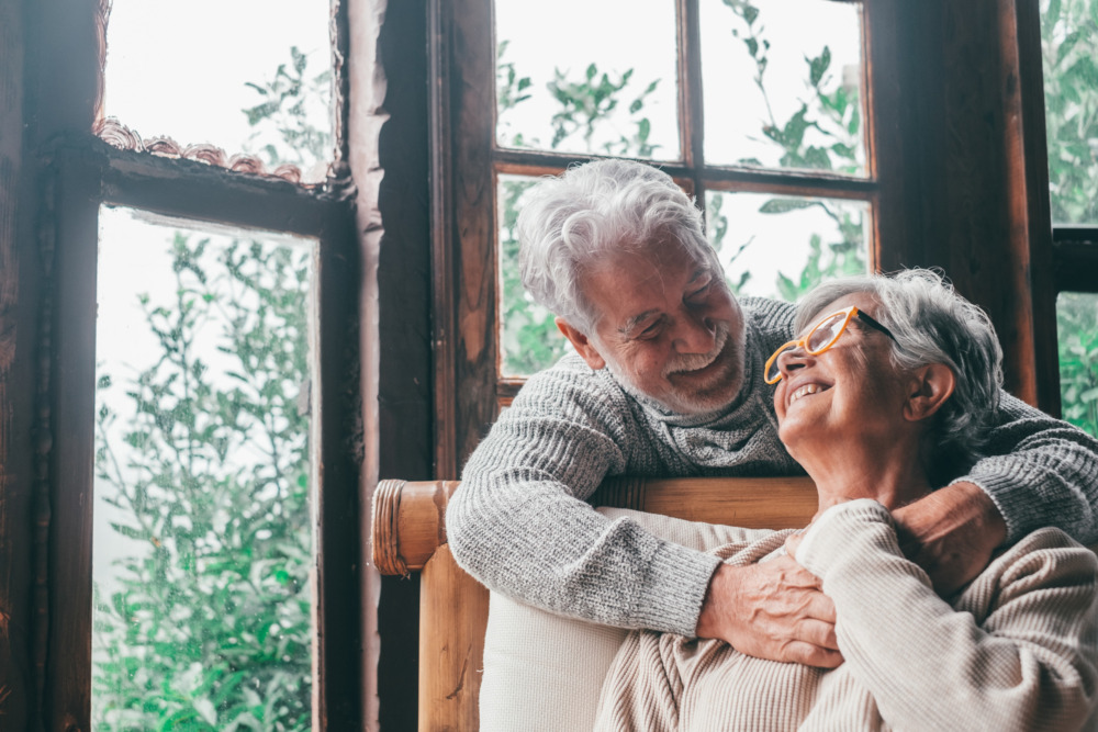 Close up image of a senior couple, the woman is sat on a comfortable chair, looking up at her husband who stands behind her with his arms around her. They smile at each other.