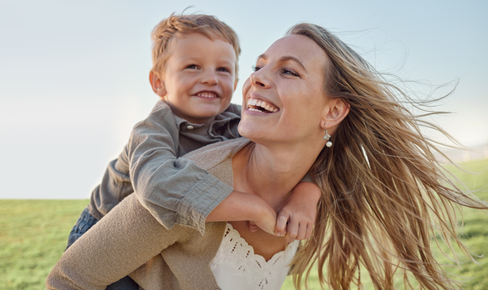 Mother giving her child a piggyback outside, both are laughing.