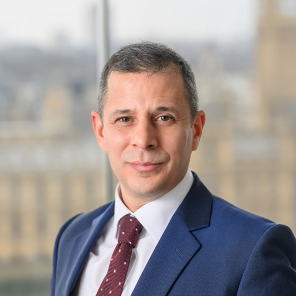 brunette man wearing navy suit with burgundy tie stood in front of houses of parliament