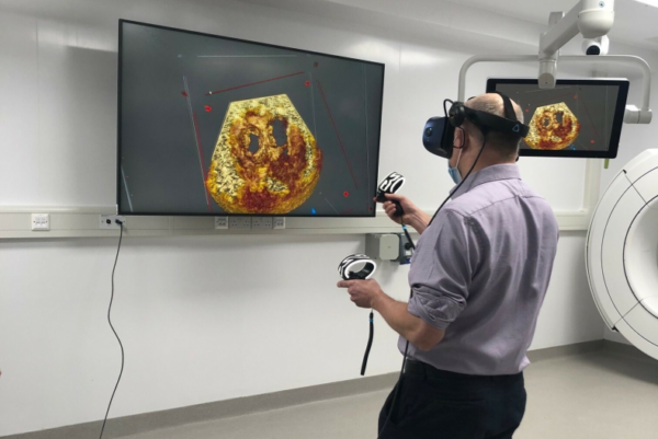 Clinician using a virtual reality headset and handheld controllers to interact with a three‑dimensional medical image displayed on a wall‑mounted screen in a clinical room.