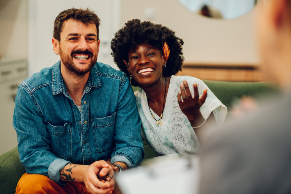 Image of a young couple smiling from over the shoulder of a consultant, who sits opposite the with their back to the camera.