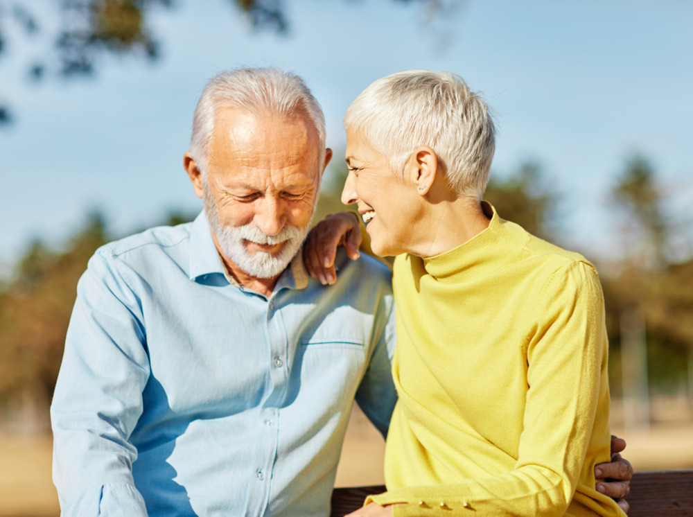 Happy senior couple sat outside enjoying the sunshine. The man looks down while the woman talks into his ear. Both are smiling.