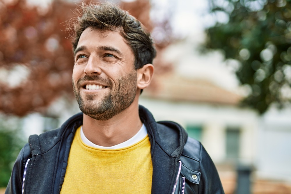 A young man facing the camera and smiling, wearing a jacket, with autumn trees behind him.