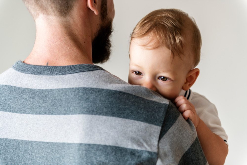 A father with his back to the camera holds his young son, who peers over his shoulder, facing us.