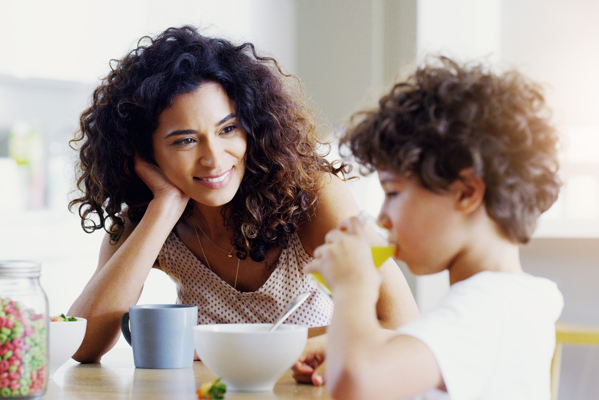 Woman with brunette, curly hair smiling at young boy drinking orange juice