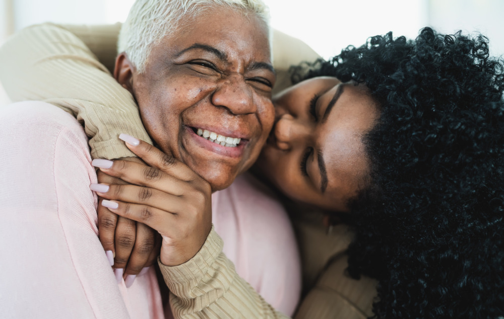 Close up image of an older woman laughing as her adult daughter hugs her from behind and kisses her cheek.