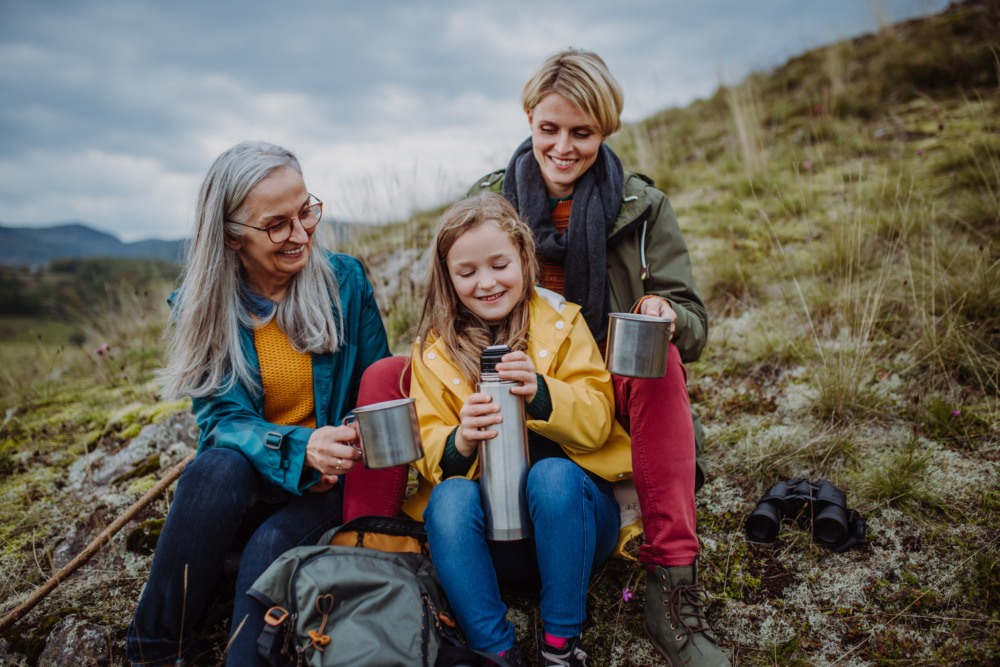 A young girl with her mother and grandmother, drinking hot tea from a flask on the side of a mountain.