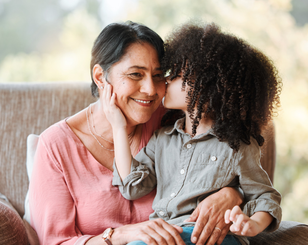 A young boy sits on his smiling grandmother's lap and kisses her cheek.