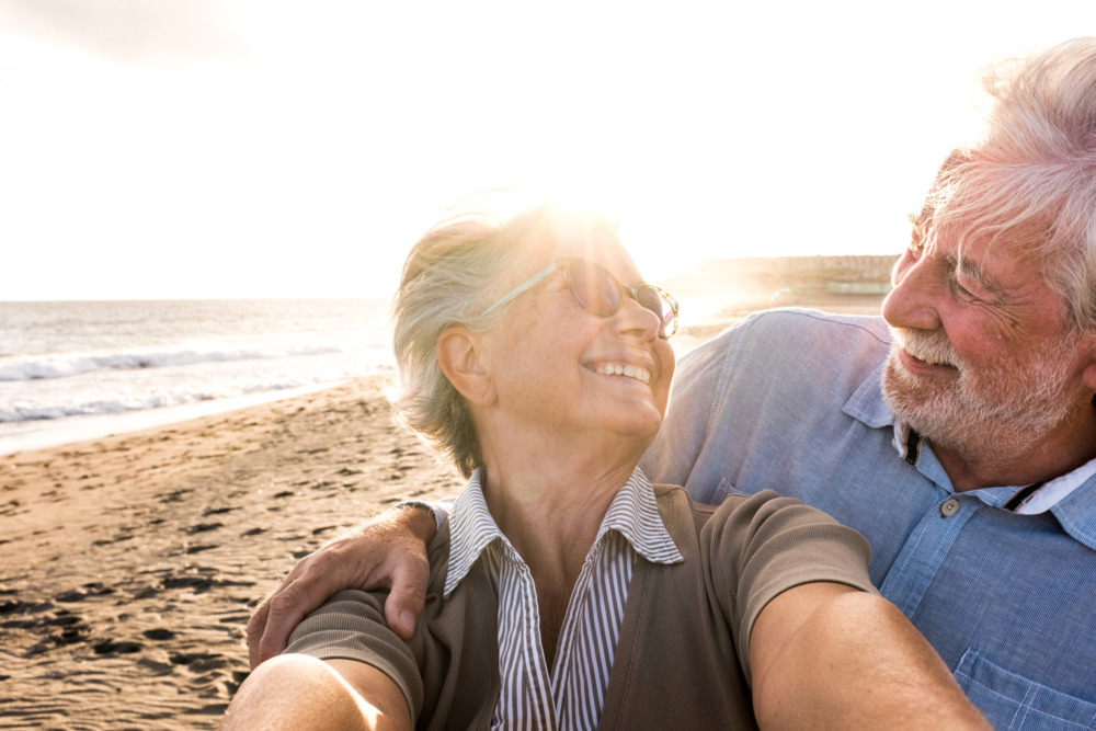 Close up image of an older couple on the beach, smiling in the sunshine. Their faces are obscured by the glare of the sun as they take a selfie together.