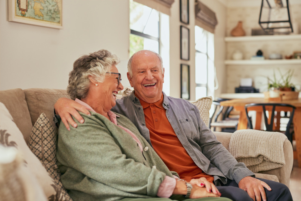 Happy retired couple sit together on a sofa, holding hands, laughing. The husband has his arm around the wife.