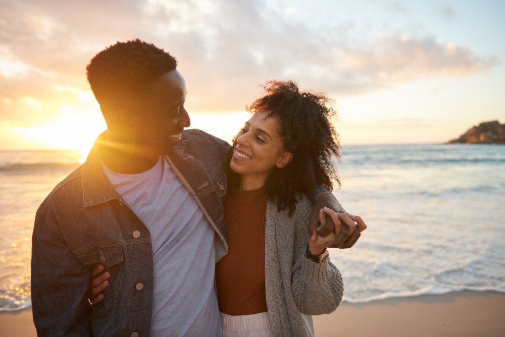 Affectionate young multiethnic couple smiling at each while standing arm in arm on a sandy beach at sunset